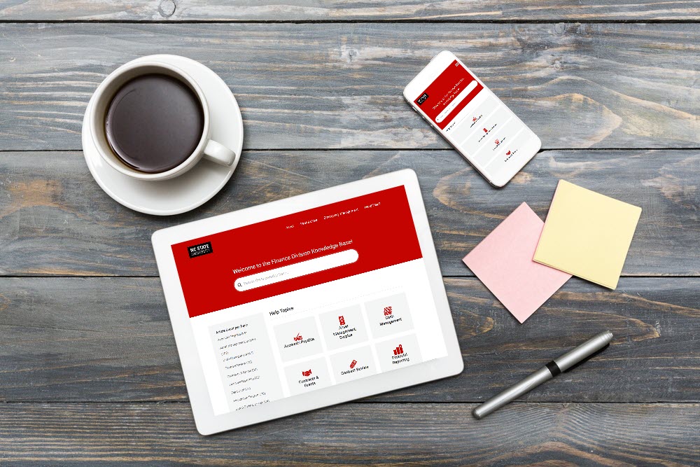 A cup of coffee, sticky notes, pen, cell phone and tablet on a wooden table. The cell phone and tablet have the Finance Division Knowledge Base open.