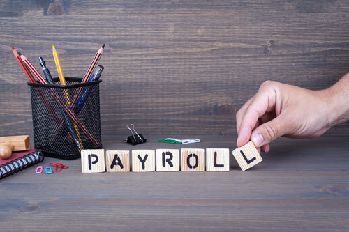 A person arranging wooden letter blocks on a desk surface to spell out PAYROLL.