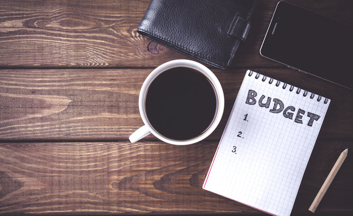 A table with a wallet, cell phone, cup of coffee, notepage and pencil on it. The word "Budget" and a numbered list are written on the notepad.