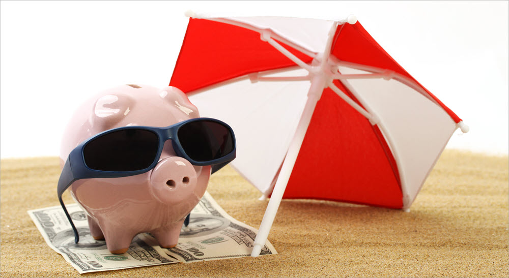 A piggy bank wearing sunglasses sits on a blanket made of money under an umbrella on the beach.