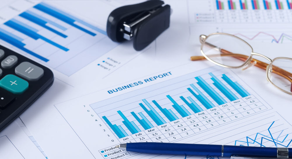 A desktop covered in papers, charts, and graphs, along with a pen, stapler, calculator, and glasses. A business report is in the center.