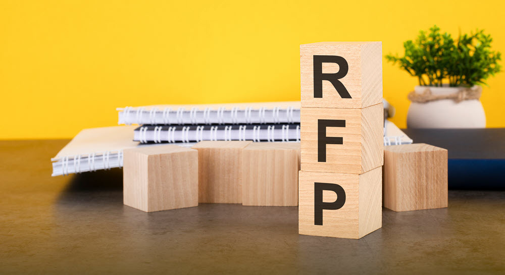 Wooden blocks sitting on a desk with notebooks. Three blocks are stacked and spell out RFP.