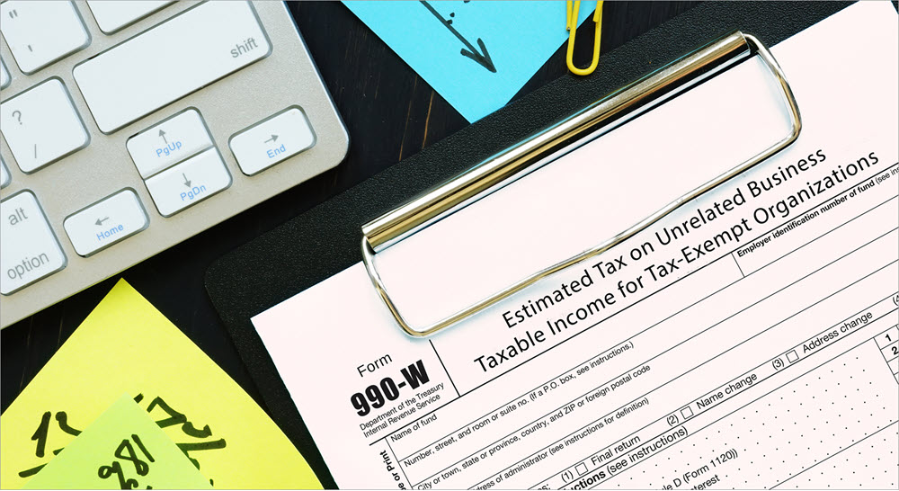 A desktop with a keyboard, paper clips, and sticky notes. On a clipboard is an IRS form 990-W.