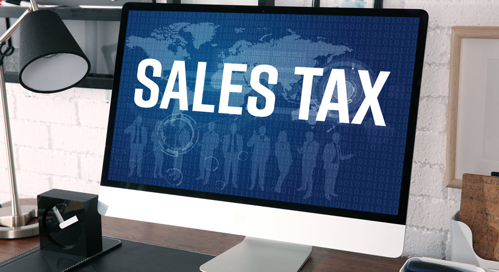 A desktop computer monitor with the words "SALES TAX" displayed on a blue background showing a world map and a group of people.