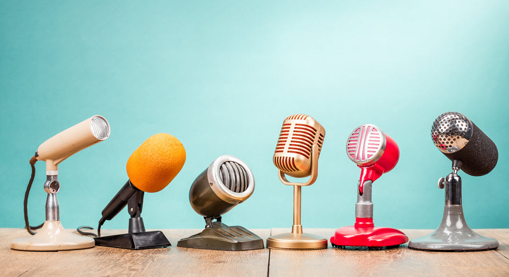 Six vintage microphones, all different styles, sitting on a wooden table
