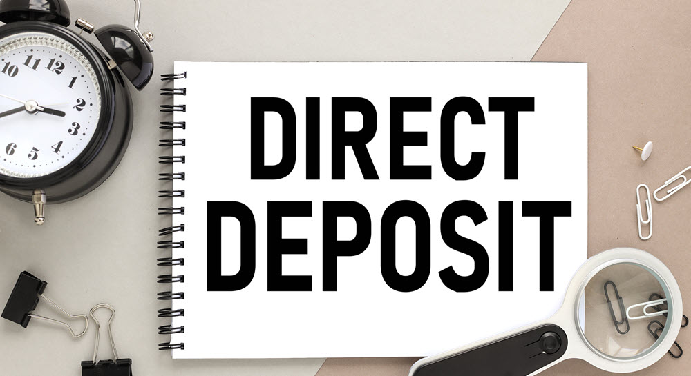 A notepad with "DIRECT DEPOSIT" written on it sitting on a desk, surrounded by a magnifying glass, an alarm clock, and paper clips.