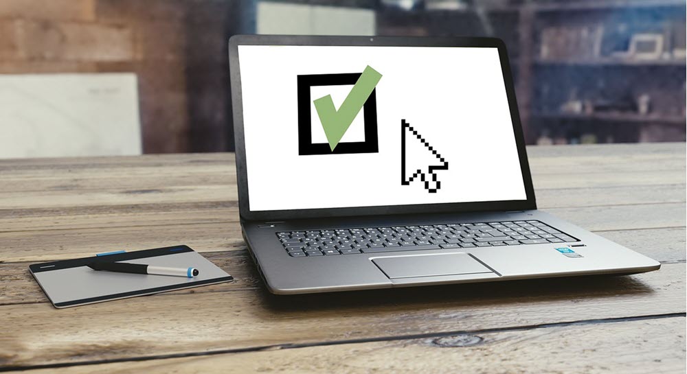 A desk with a laptop and writing pad sitting on it. The laptop shows a large mouse cursor and a large checkbox with a green checkmark