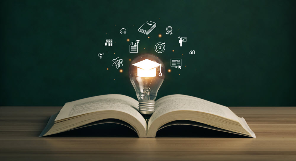 A book open on a desk with a lightbulb in the middle surrounded by icons symbolozing learning and knowledge