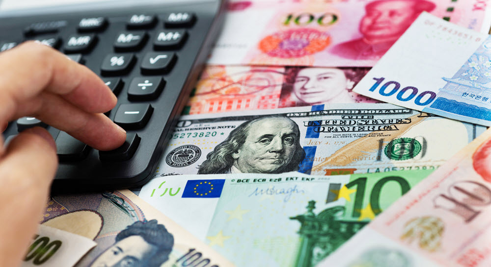 A person's hand using a calculator that is resting on a desk covered in various bills in various currencies