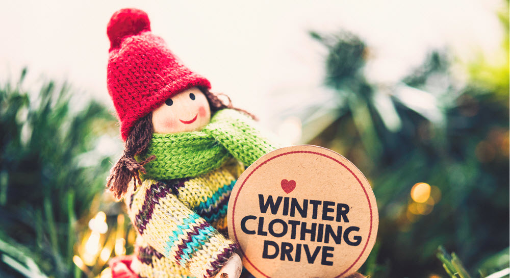 A wooden doll wearing winter clothing holding a sign saying "Winter Clothing Drive"