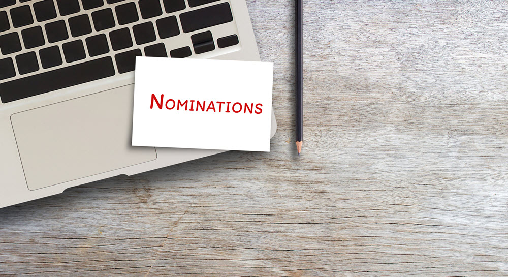 A laptop computer on a table. A card reading "Nominations" is resting on the computer.
