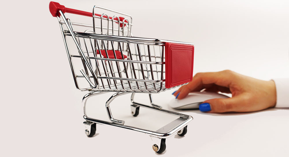 A small metal shopping cart resting on a table in front of a hand using a computer mouse