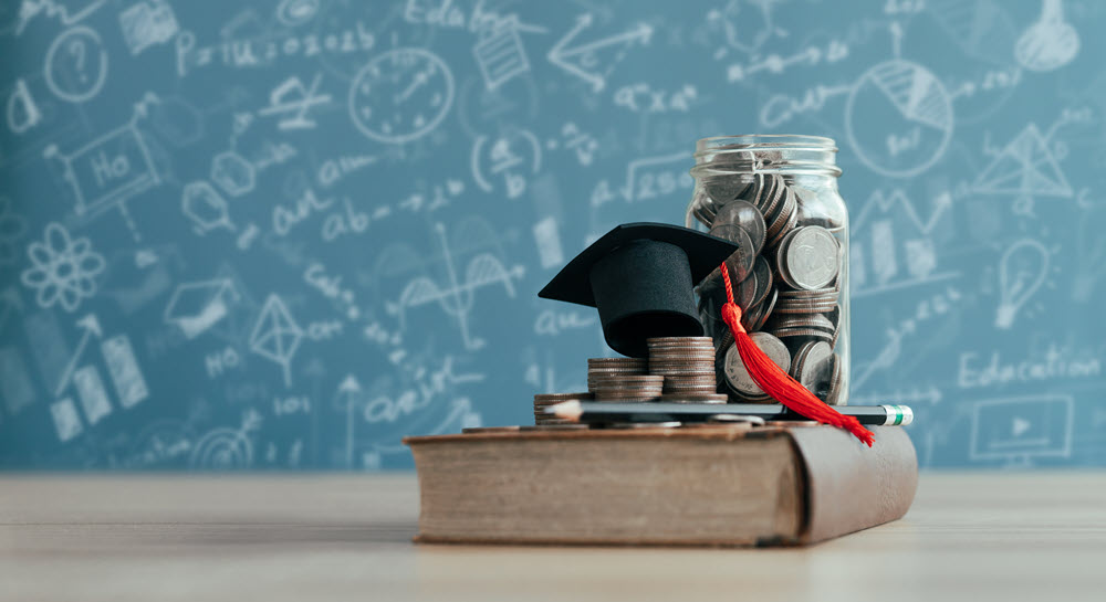 A jar of coins, a graduation cap and a pencil on a large book with a blackboard in the background.